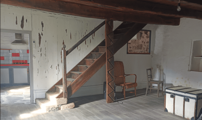Image du salon actuel dans une vieille maison en pierres, murs défraichis, sol en vinyle gris, mur en chaux blanche, plafond en bois sombre, petites pièces séparées par des cloisons bois